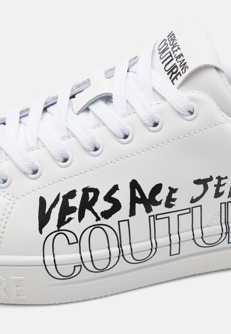 Zapatilla blanca con cuero texturizado, punta redonda y cordones planos. Presenta letras negras prominentes "VERSACE JEANS COUTURE" en el costado.