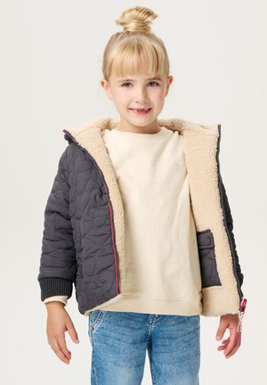 Gequilted donkergrijze jas met een beige faux fur voering. Voorzien van een ritssluiting aan de voorkant, geribde manchetten en een kleurrijke ritssluiting. G gedragen over een lichte sweatshirt.