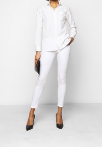 Camicia bianca con maniche lunghe e una tasca, abbinata a pantaloni bianchi attillati e scarpe con tacco nere, mentre tiene una borsa a mano nera.