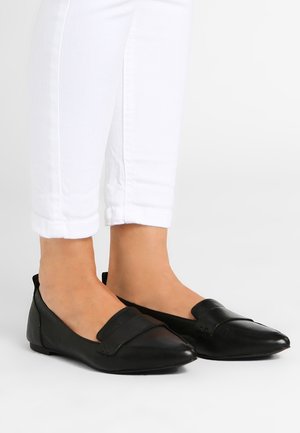 Pieds portant des mocassins en cuir noirs à bout pointu associés à un pantalon blanc raccourci sur un fond clair uni.