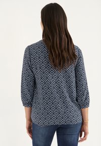 Bluse mit blauem Muster und geometrischem Design, runder Halsauschnitt und drei Viertelärmeln, kombiniert mit dunkelblauen Jeans.