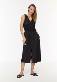 Vestido negro sin mangas con escote abotonado, cintura elástica, bolsillos laterales y una abertura a la altura de la rodilla. Llevado con sandalias negras.