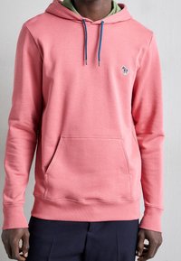 Homme portant un sweat à capuche rose avec des cordons bleus et un petit patch zèbre multicolore sur la poitrine, assorti à un pantalon foncé.