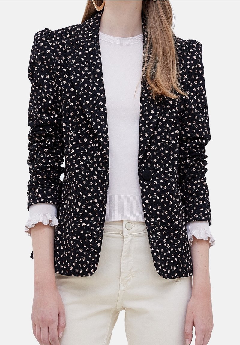 Femme portant un blazer noir à petit motif floral avec des épaules bouffantes, une chemise blanche à manches longues avec des poignets à volants, et un pantalon beige.