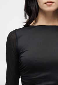 Γυναίκα από τους ώμους έως τα χείλη φοράει εφαρμοστή μαύρη μπλούζα με διαφανή μακριά μανίκια και λαιμόκοψη σκάφος, μπροστά σε απλό φόντο.