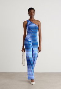 En axelbandslös jumpsuit i blå och vita ränder; åtsittande överdel, raka ben, slätt tyg; bärs med en lätt axelväska och klackar.