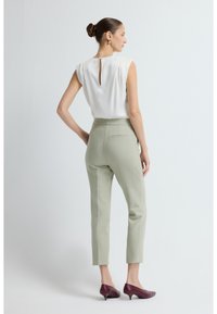 Vrouw met een knot in het haar, gekleed in een mouwloos wit blouse, lichtgroene op maat gemaakte broek en bordeauxrode schoenen met een lage hak, tegen een effen achtergrond.