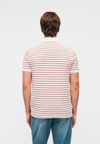 Polo blanc à manches courtes avec des rayures rouges horizontales, doté d'un col classique et d'un ourlet droit. Porté avec un jean bleu.