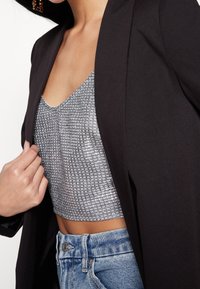 Svart blazer över en skirrande silverfärgad kort topp med rutmönster, tillsammans med högmidjade ljusblå jeans.