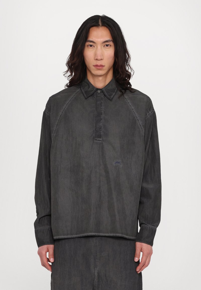 Chemise grise à manches longues avec col, patte de boutonnage et manches raglan. Présente des coutures visibles et un petit logo sur la poitrine.