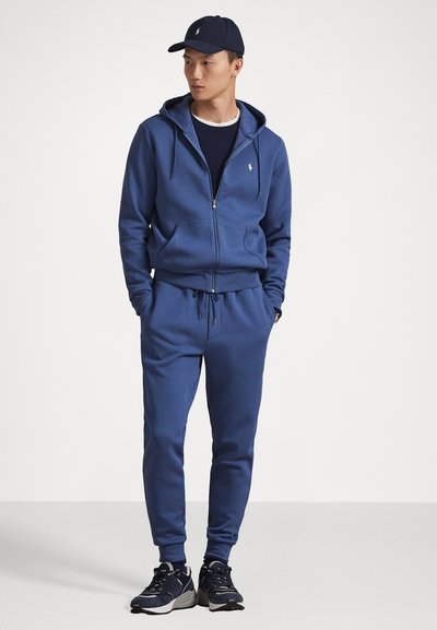 Zila ar rāvējslēdzēju aizdare, ar auklu, saskaņoti treniņbikses un tumši zila cepure. Materiāls izskatās mīksts ar gludu tekstūru. Smalks logo uz krūtīm.