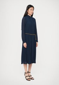 Γυναίκα φορούσε ένα ναυτικό μπλε μακρυμάνικο midi φόρεμα με πτυχώσεις και μια λεπτή χρυσή αλυσίδα, συνδυασμένο με μαύρα ψηλοτάκουνα σανδάλια με λουριά.