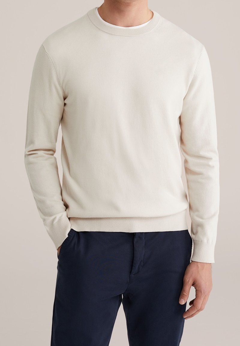 Homme portant un pull uni beige clair à col rond avec manches longues et un pantalon bleu marine foncé, debout avec une main dans la poche.