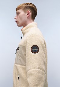 Fleece jacket in cream with a contrasting beige panel. Features a zip-up collar, chest pocket, and a round logo patch on the sleeve.