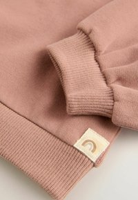 Sweatshirt rose clair avec des poignets et un ourlet côtelés. Présente une étiquette en tissu avec un design arc-en-ciel. Texture douce, design décontracté.
