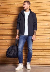 Černá lehká bunda, bílý tričko, modré džíny a bílé tenisky. Drží černou tašku přes rameno. Stojí u dřevěné zdi.