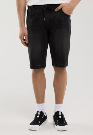 Schwarze Denim-Shorts mit einem Kordelzugbund, Seitentaschen und geradem Schnitt. Kombiniert mit schwarzen Turnschuhen und weißen Socken.