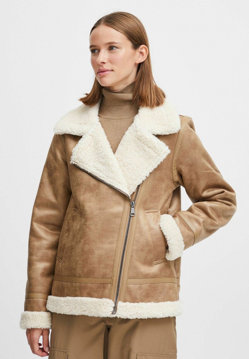 Veste en peau de mouton brune avec un col et des poignets en fausse fourrure crème, dotée d'une fermeture éclair devant et de poches latérales. Texture lisse avec une coupe structurée.