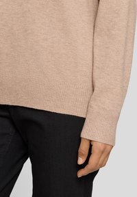 Beiger Strickpullover mit geripptem Saum und Bündchen, mit entspanntem Schnitt. Kombiniert mit schwarzen Hosen, um die Textur des Stoffs zu betonen.