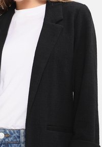 Černé texturované sako s jedním knoflíkem a klopovým límcem, nošené přes jednoduchou bílou košili. Má dvě boční kapsy.
