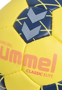 Gelb und marineblaues Hummel Classic Elite Handball mit weißen Chevron-Logos und pinkem Markentext, der auf den Paneelen sichtbar ist.