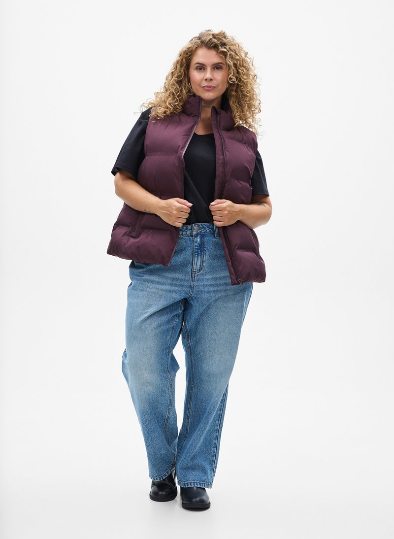 Kvinde med krøllet hår iført en lilla vatteret vest, sort t-shirt, løse blå jeans og sorte sko, stående foran en ensfarvet hvid baggrund.