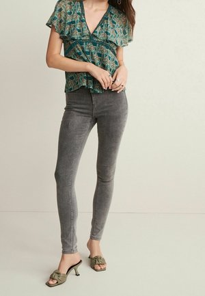 Kobieta ubrana w zieloną bluzkę z dekoltem w serek w wzory, z falbankami na rękawach, szare obcisłe jeansy oraz oliwkowe sandały na obcasie z kokardką.