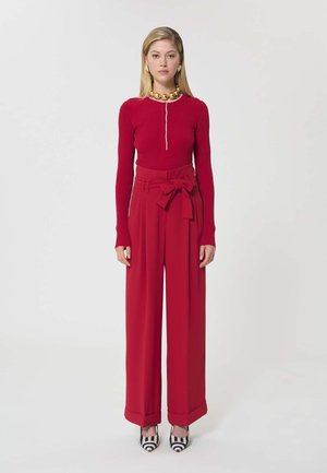 Femme debout portant un haut rouge à manches longues côtelé, un pantalon large taille haute rouge avec une ceinture à nœud, un collier en or et des talons à motifs noir et blanc.