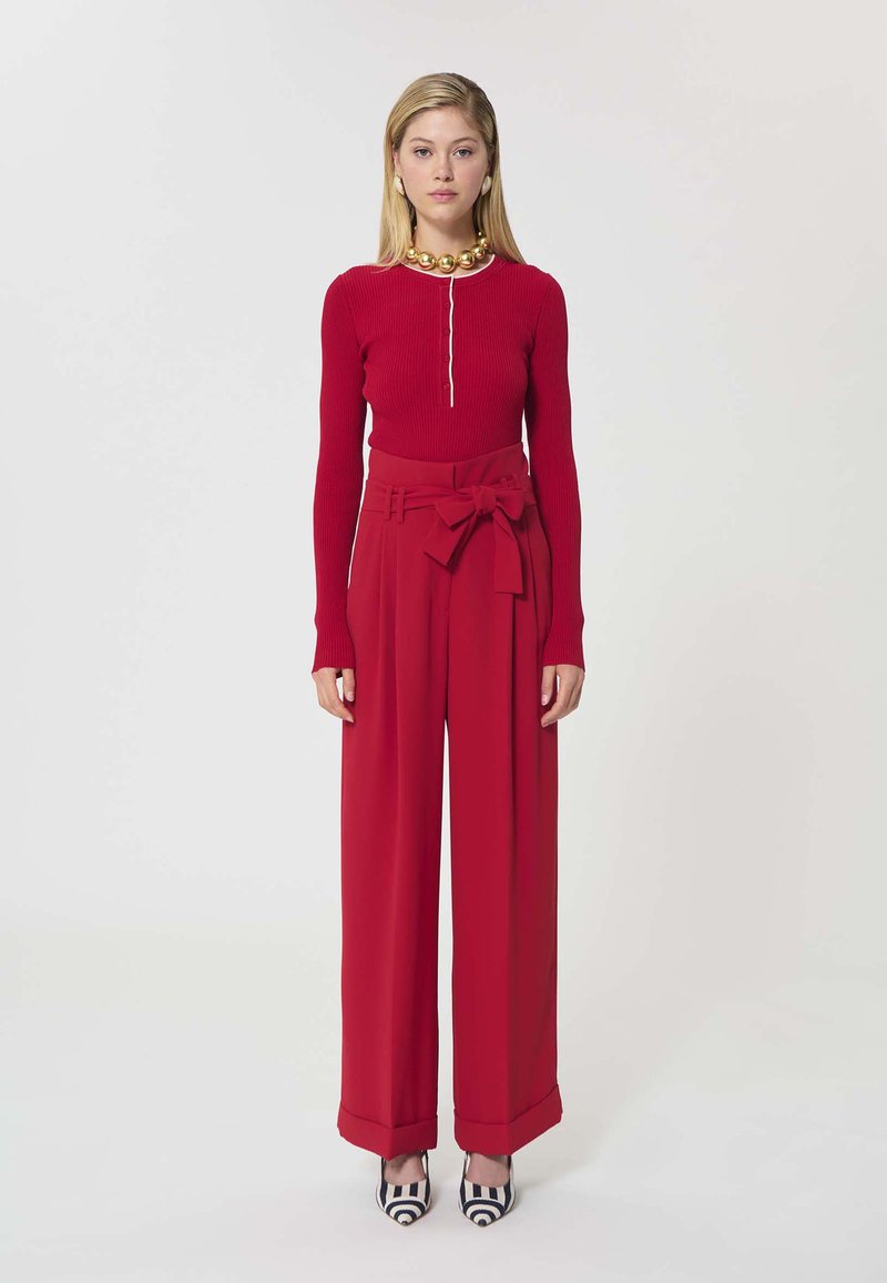 Femme debout portant un haut rouge à manches longues côtelé, un pantalon large taille haute rouge avec une ceinture à nœud, un collier en or et des talons à motifs noir et blanc.