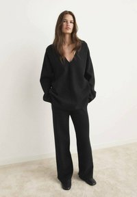 Schwarzer, übergroßer Pullover mit tiefem V-Ausschnitt, kombiniert mit weit geschnittenen schwarzen Hosen. Weiche Textur, minimalistisches Design. Flache schwarze Schuhe.