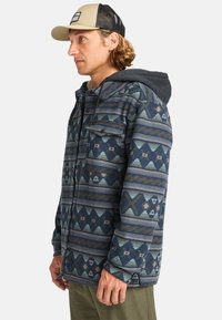 Textured blue and gray patterned jacket with geometric designs, two chest pockets, and a gray hood. Underneath, a light gray shirt is visible.