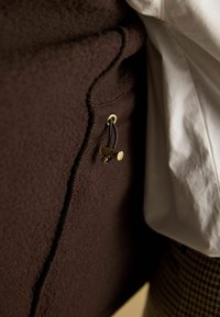 Brązowa tkanina z widocznym szwem i małym złotym zapięciem, obok białego rękawa i odzieży w wzór w houndstooth.