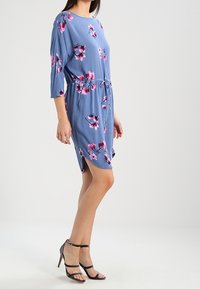 Sininen kukallinen mekko, jossa on 3/4-hihat, vyö solmimiseksi ja sivuhalkiot, pehmeällä pinnalla ja vaaleanpunaisella kukkakuviolla. Yhdistetty mustiin remmisandaaleihin.