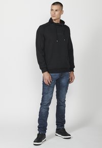 Sudadera negra con un patrón texturizado, capucha con cordones y puños acanalados, combinada con jeans cargo azules y zapatillas negras con acentos blancos.