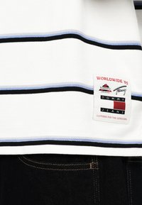 Λευκό πουκάμισο κοντομάνικο με μαύρες και ανοιχτό μπλε οριζόντιες ρίγες, διαθέτοντας ένα λογότυπο στη χαμηλή πλευρά.