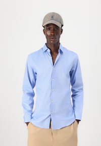 Camisa de botones de color azul claro con mangas largas, cuello y una textura sutil; combinada con una gorra gris que presenta un pequeño logotipo.