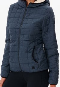 Marineblå quilted jakke med hætte, der har lynlåslukning, sidelommer med sort kant og en blød indvendig foring.