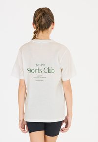 T-shirt couleur crème à manches courtes avec un imprimé vert au dos portant la mention "Les’Arcs Sports Club Luxury Athleisure Wear."