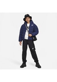 Giacca puffer navy con cappuccio, felpa nera con logo Nike, pantaloni neri a sigaretta, sneakers nere e cappello a secchiello nero.