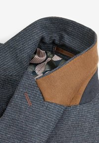 Villane segu blazer, mille tekstuuriga sinise välispinnaga, pruunast nahast kraega ja mustrilise voodriga, millel on rohelised ja roosad tooni.