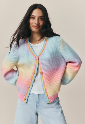 Junge Frau mit langen dunklen Haaren trägt eine pastellfarbene Regenbogenstrickjacke über einem weißen Hemd und hellblauen Jeans, stehend vor einem schlichten Hintergrund.