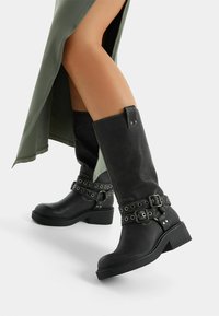 Botas negras hasta la rodilla hechas de cuero texturizado con múltiples hebillas y tachuelas metálicas circulares, con un tacón grueso para mayor estabilidad.