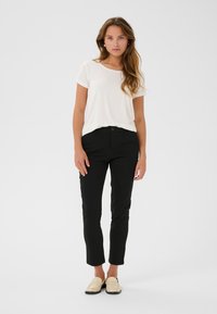 Černé slim-fit kalhoty s pasem na knoflík, v kombinaci s bílým tričkem s krátkým rukávem. Obuv tvoří béžové mokasíny. Jednoduchý design a hladká textura.