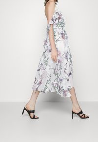 Blommig midi-klänning med volangärmar utan axlar, tillverkad av vitt tyg prytt med lila och grönt botaniskt mönster. Bärs med svarta remsandaler.