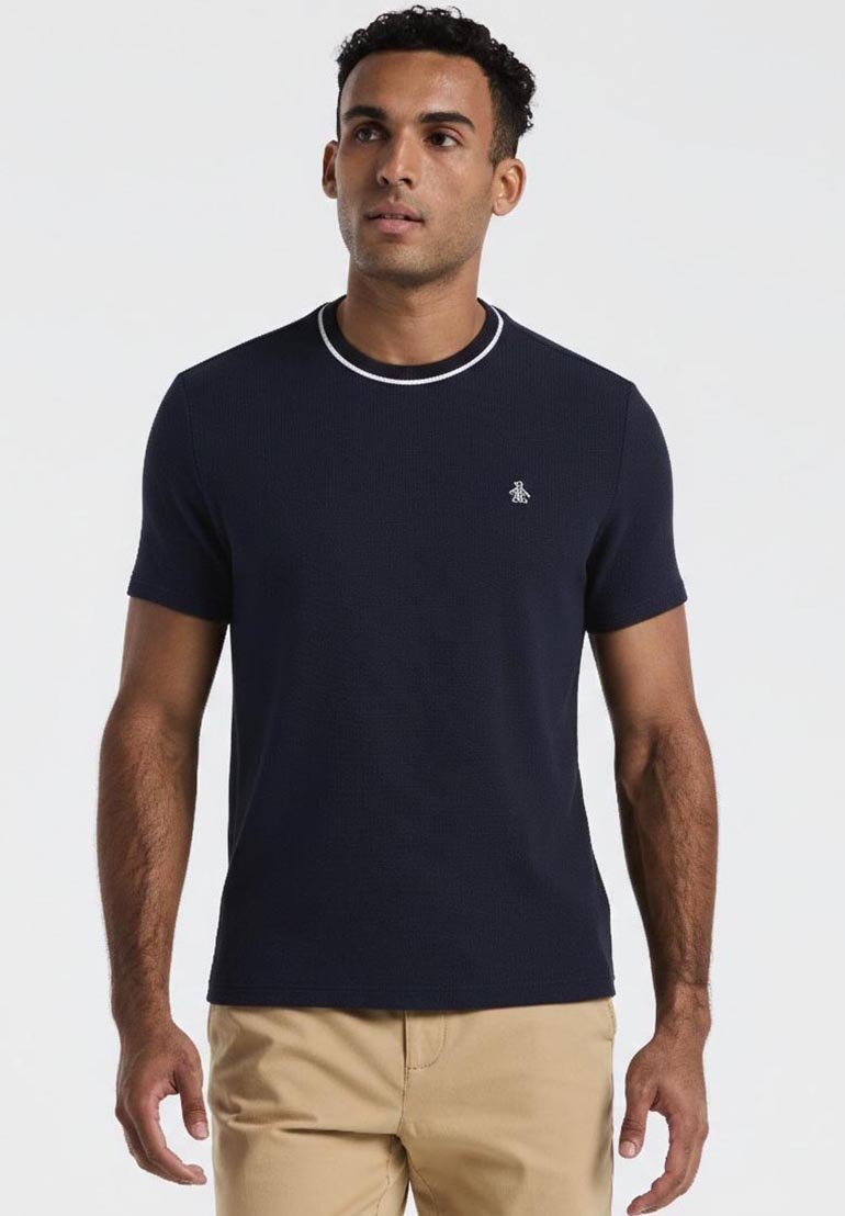 Homme portant une chemise bleu marine à manches courtes avec un bord blanc sur le col et un pantalon kaki clair, debout devant un fond uni.