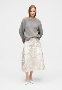 Pilkas megztinis, metalinis midi sijonas su gėlių raštais, balti smailūs bateliai. Modelis stovi sukryžiavęs rankas priešais vientisą foną.