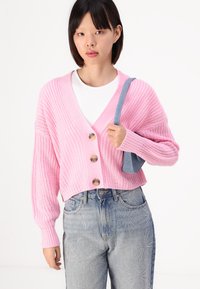 Rosa strikket cardigan med V-hals, tre store knapper og brede ermer. Kombinert med høytlivs denim jeans med dekorativ stikning.