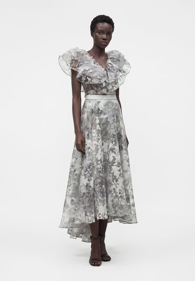 Γυναίκα που φοράει αμάνικο γκρι φόρεμα με λουλουδένιο σχέδιο, με στρωμένο βολάν στο ντεκολτέ και ασύμμετρο τελείωμα, στέκεται μπροστά σε λευκό φόντο.