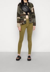Takki, jossa on camouflage-kuvio vihreässä ja vaaleassa beessä, musta vaippalaukku, oliivinvihreät kapeat farkut ja mustat korkokengät. Pintakuvio on sileä koko asetelman ajan.
