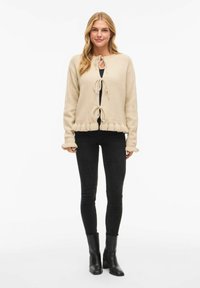 Beige strikket cardigan med rund hals, knyting foran og rysjekant på ermene. Brukt over en svart topp og kombinert med svarte smale jeans.
